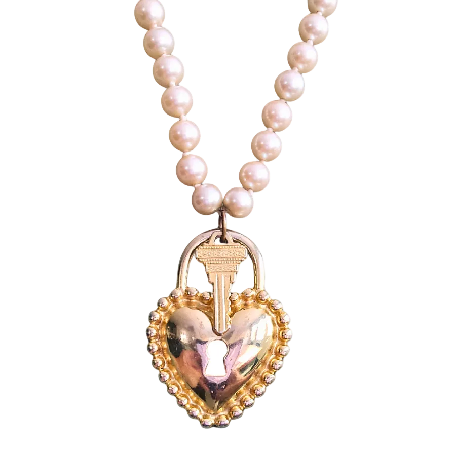 A pearl necklace with a heart-shaped lock pendant, featuring a gold heart frame, a key in the lock, and a pink heart-shaped mirror in the center, set against a plain white background.