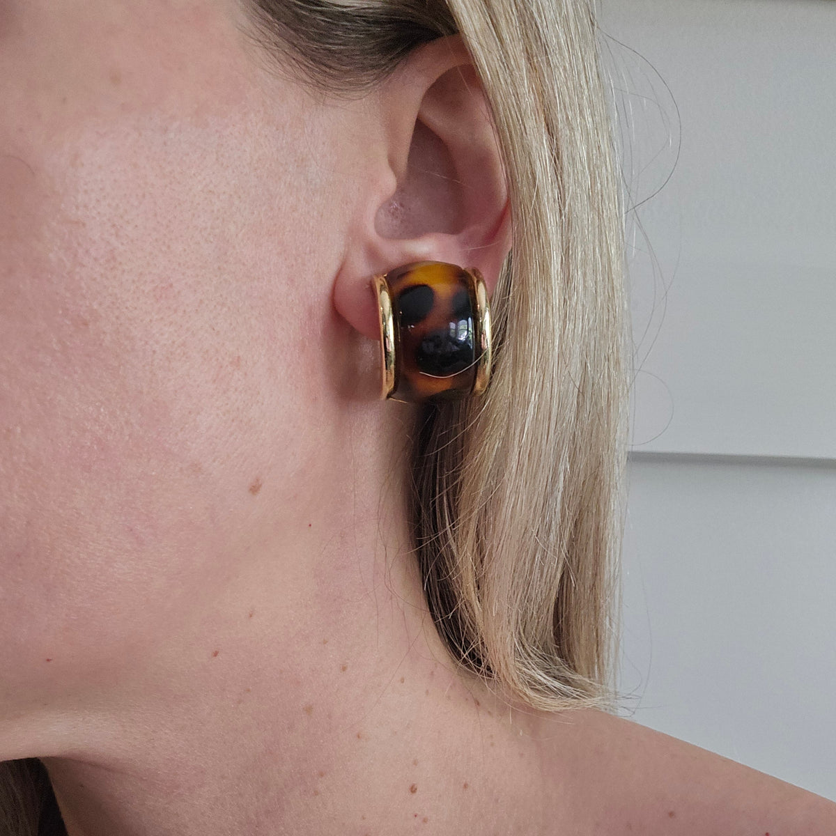 A close-up of a model's ear wearing a chunky, square-shaped hoop earring with a glossy tortoiseshell pattern and gold-toned metal borders.