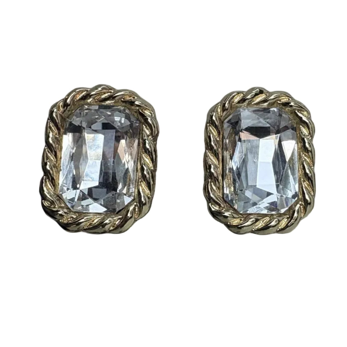 A pair of gold-toned statement earrings with large clear rectangular faceted crystals, displayed on a textured stand against a light gray fabric backdrop.