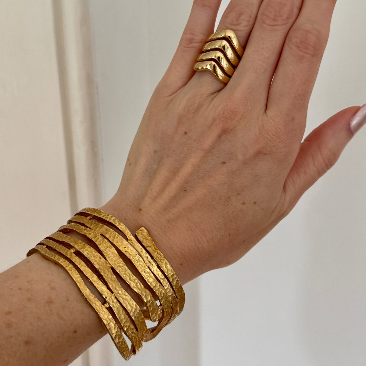 A close-up of a person's hand and wrist wearing textured gold jewelry, including a multi-band cuff bracelet on the wrist and a wavy multi-band ring on the finger, against a plain off-white background.