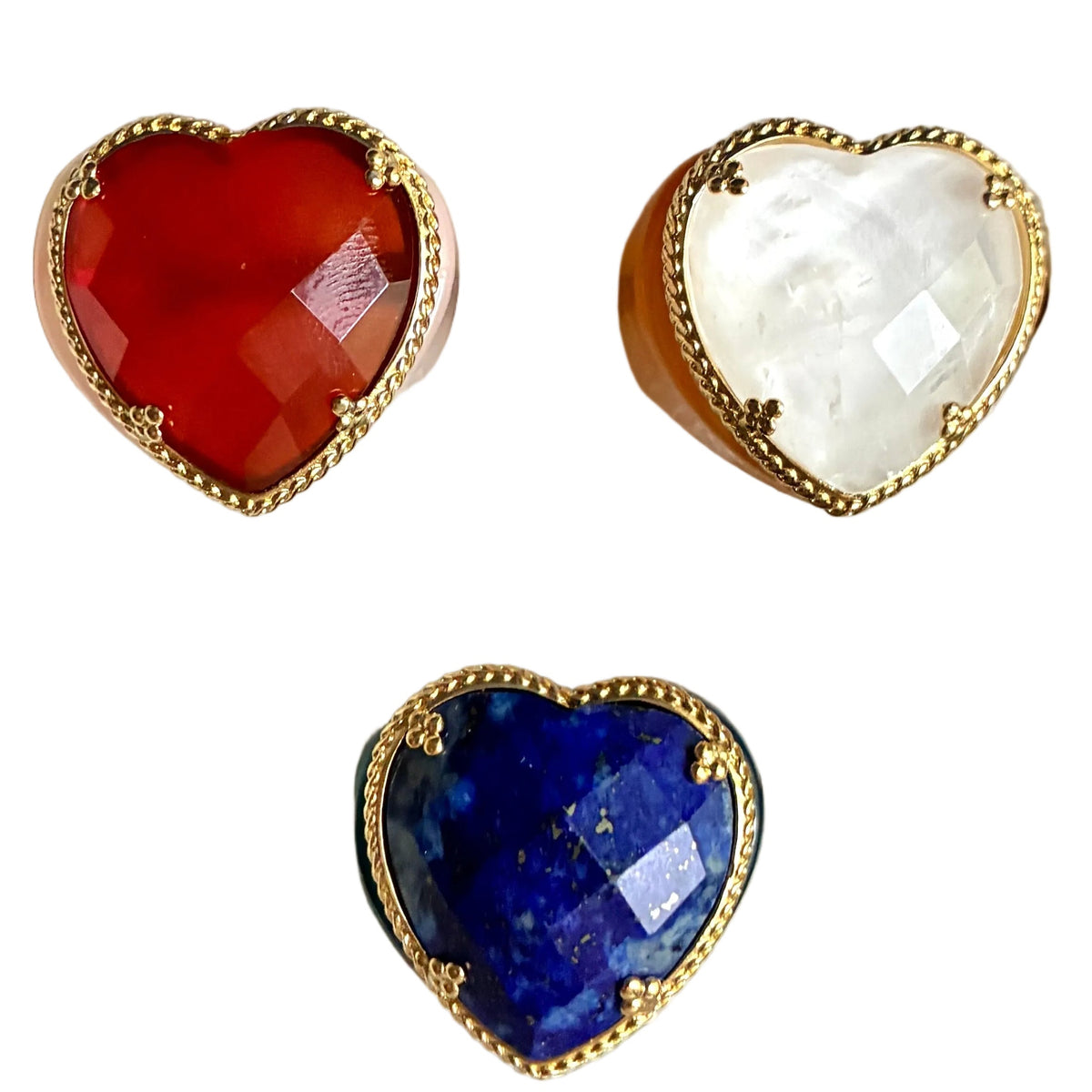 Three heart-shaped ring toppers in red, white, and blue stone settings with gold-tone borders, displayed on a cream fabric ring box.