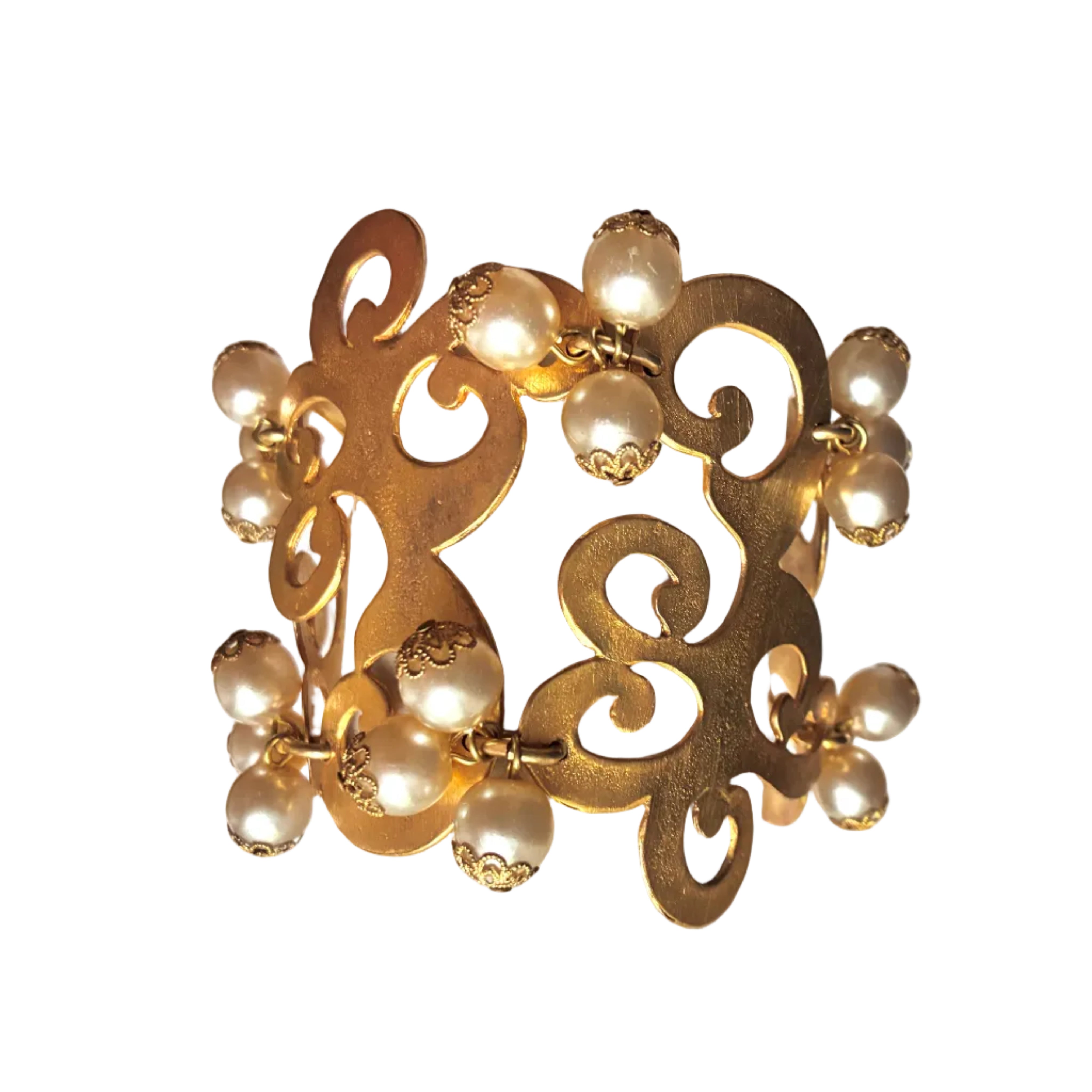A wide gold-tone cuff bracelet with an ornate openwork scroll design and multiple dangling pearl-like beads, displayed on a white cylindrical jewelry stand.