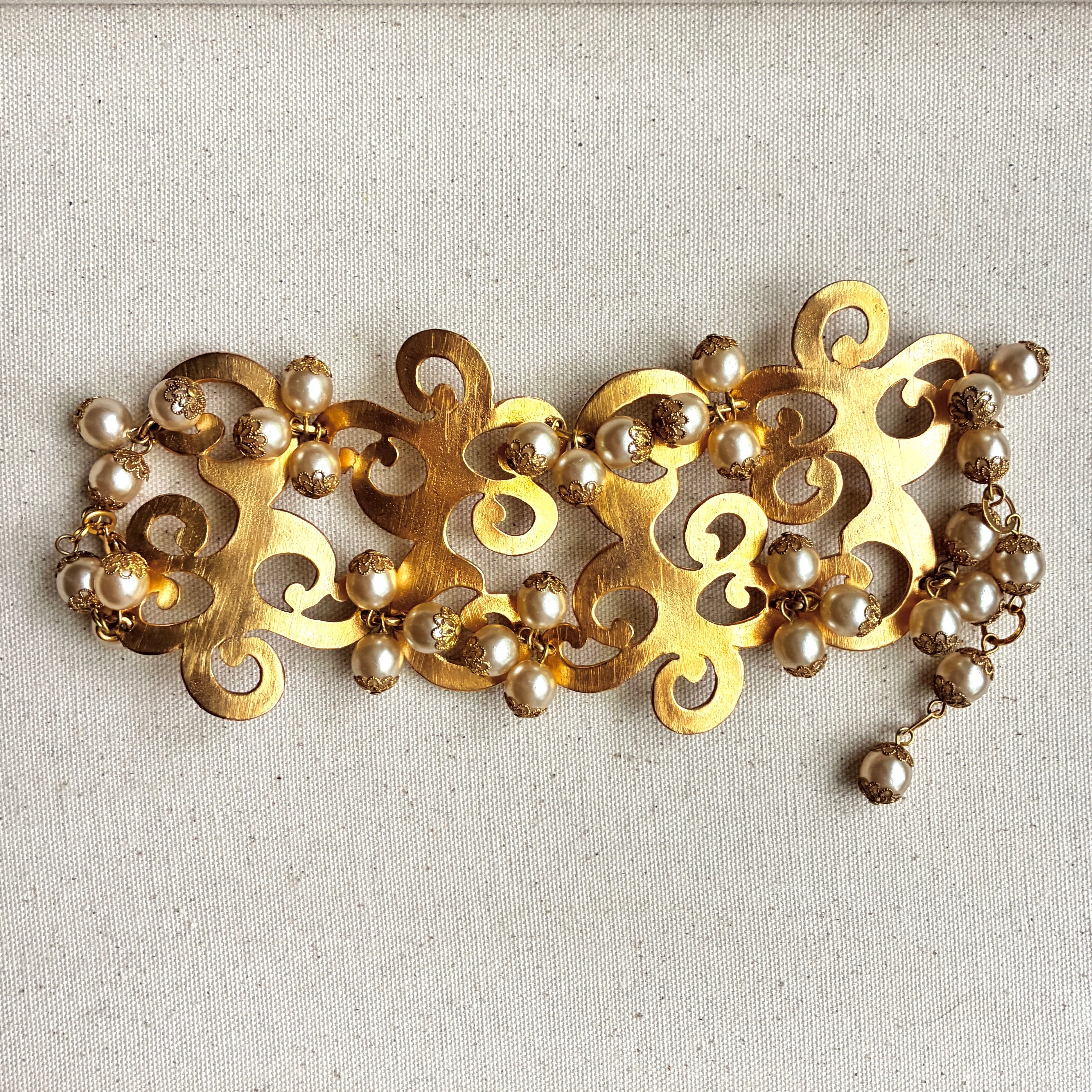 A vintage-style gold filigree cuff embellished with small pearl-like beads, resting on a textured beige fabric surface.