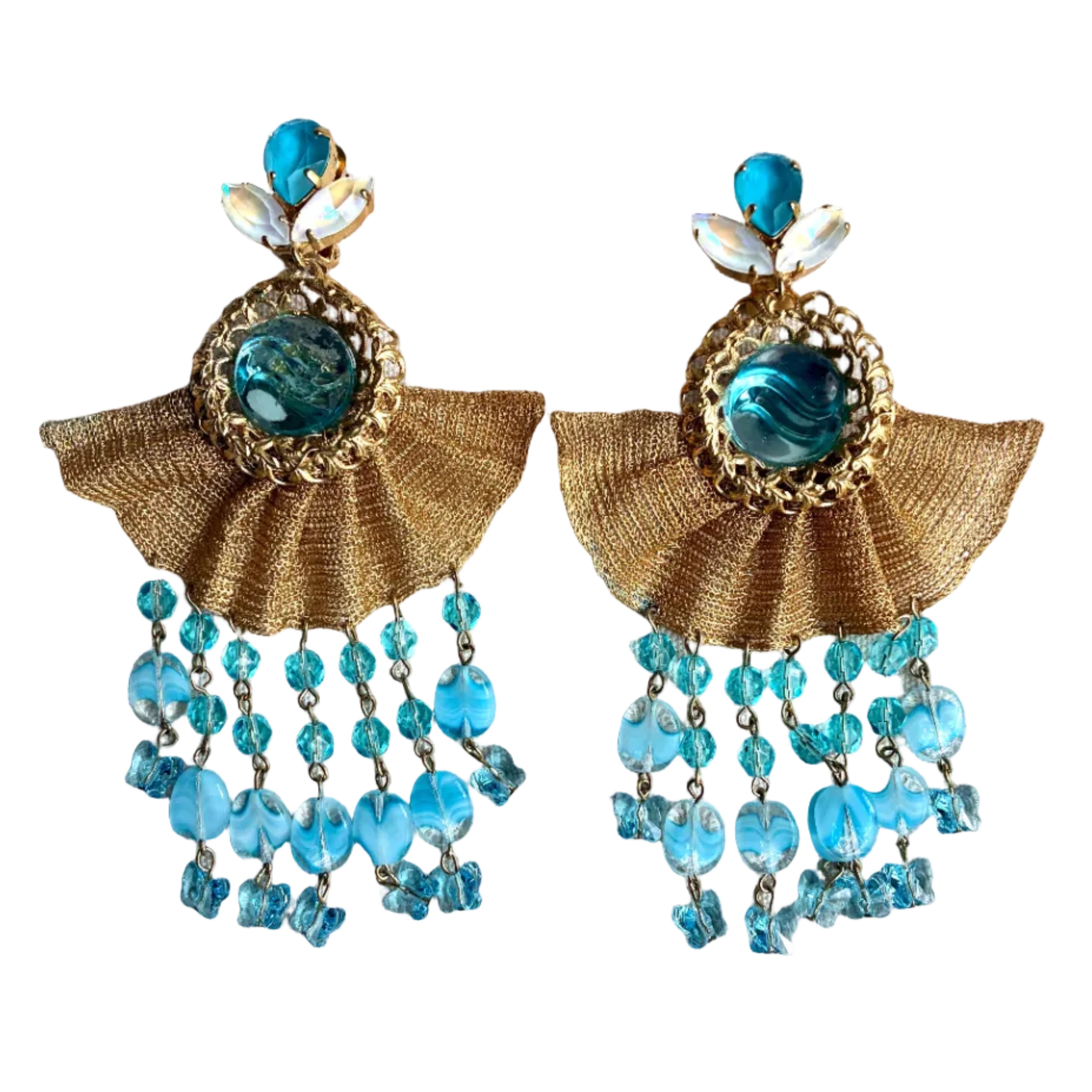 A pair of gold and blue statement dangle earrings displayed on a textured beige fabric surface. The earrings feature a gold-tone fan-shaped frame with a central blue faceted stone, white shell accents at the top, and cascading rows of light blue beads and crystal drops.