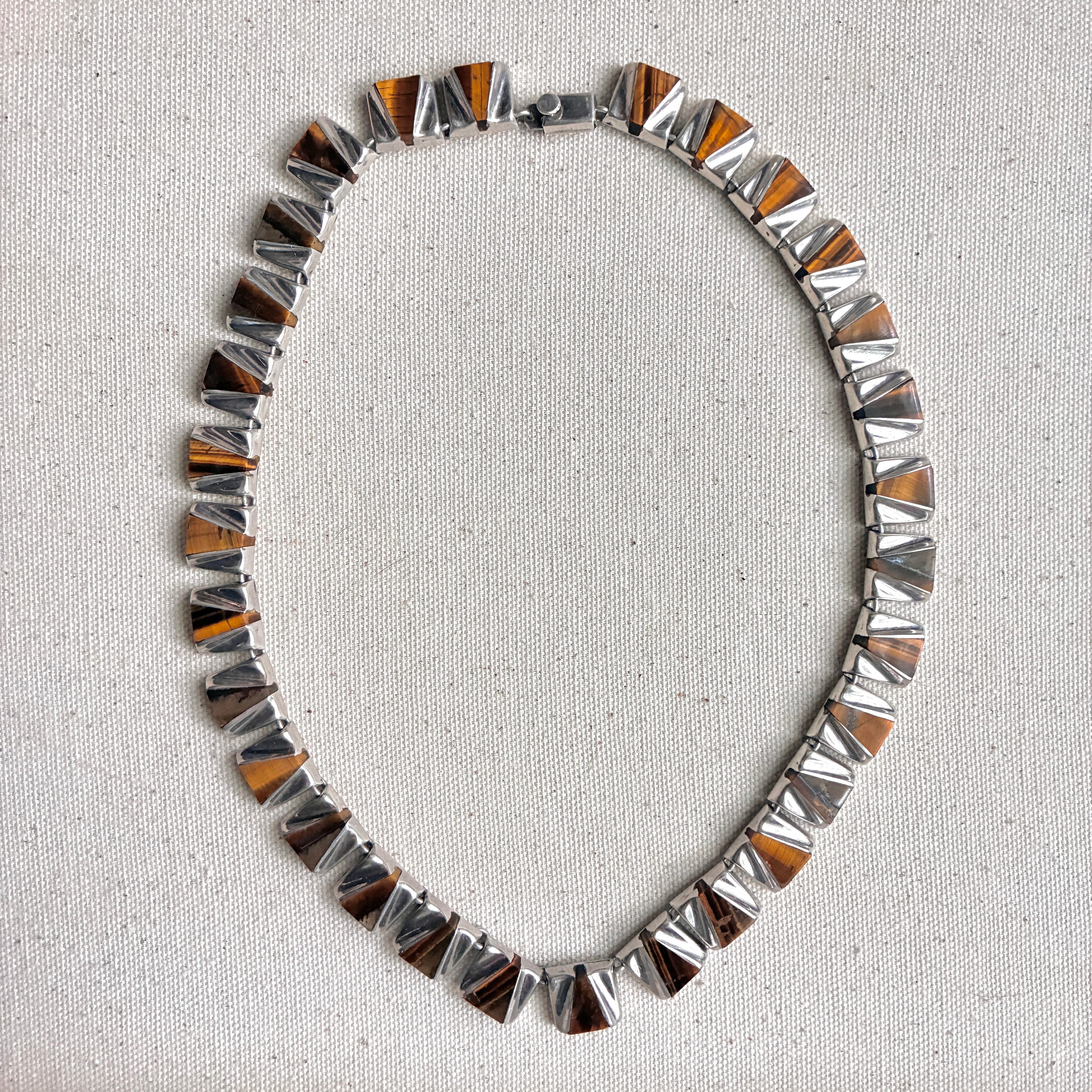 A silver-toned geometric link chain necklace with alternating triangular segments featuring orange and dark brown tigers eye details, displayed on a textured off-white fabric surface.