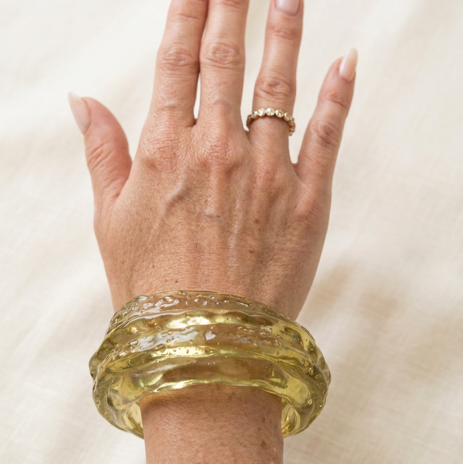 A close-up of a person's hand and wrist wearing a chunky, textured translucent green bangle bracelet, with a delicate gold ring on the middle finger, set against a soft beige fabric background.