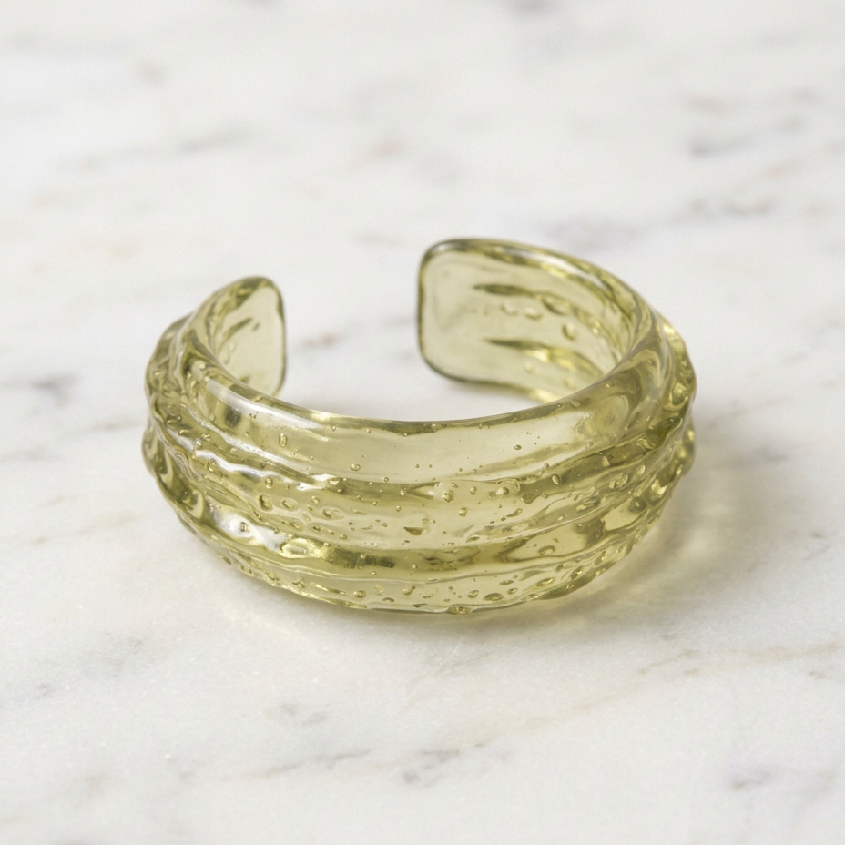 A textured, hammered glass cuff in a translucent pale yellow-green color, resting on a white marble surface.