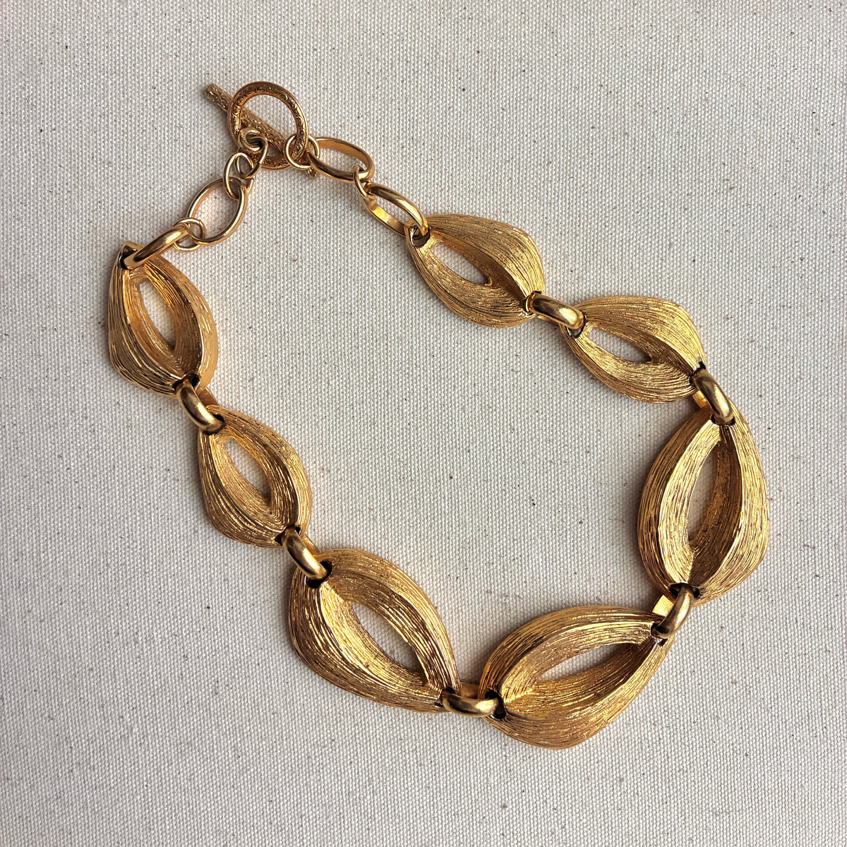 A gold link necklace with textured, leaf-like oval links, displayed on a beige fabric jewelry box insert.