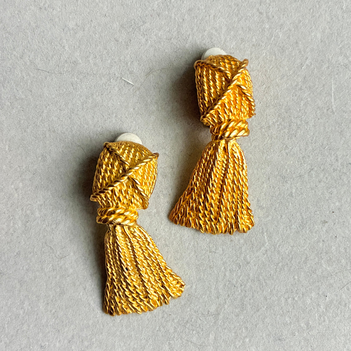 A pair of gold tassel earrings with a textured, braided thread design and white backing, displayed on a neutral gray background.
