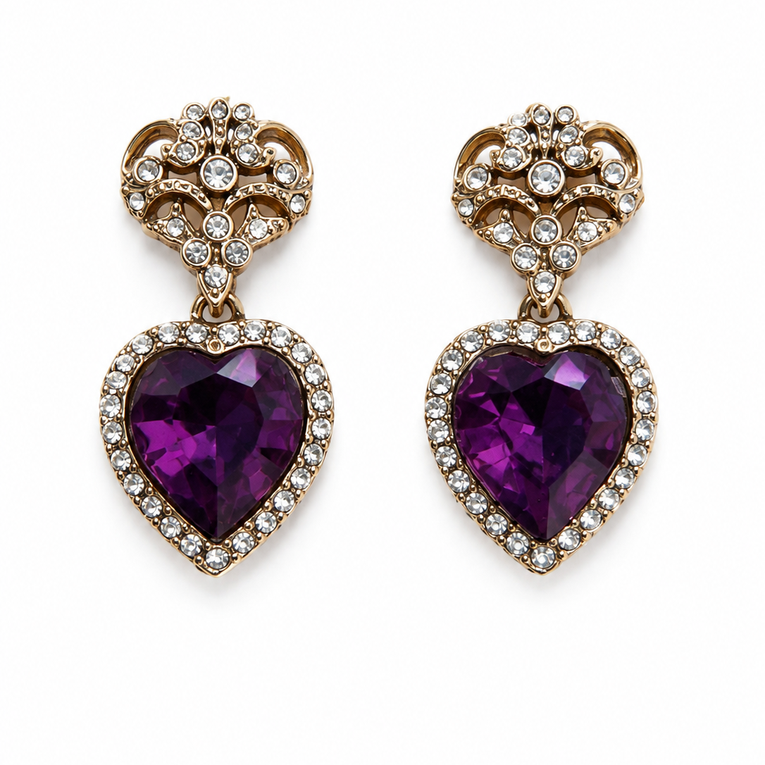 A pair of gold heart-shaped drop earrings featuring large purple heart-shaped gemstones surrounded by small clear stones, set against a plain white background.