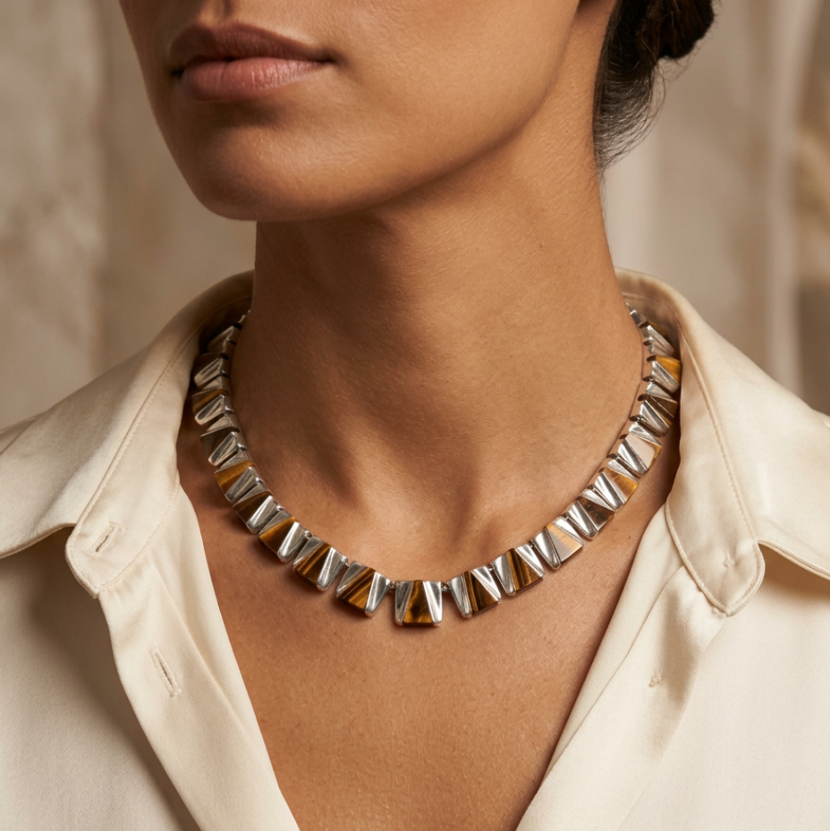 A close-up of a model wearing a chunky silver geometric beaded necklace with alternating triangular and rectangular links, geniune tigers eye styled with a cream-colored collared shirt. 