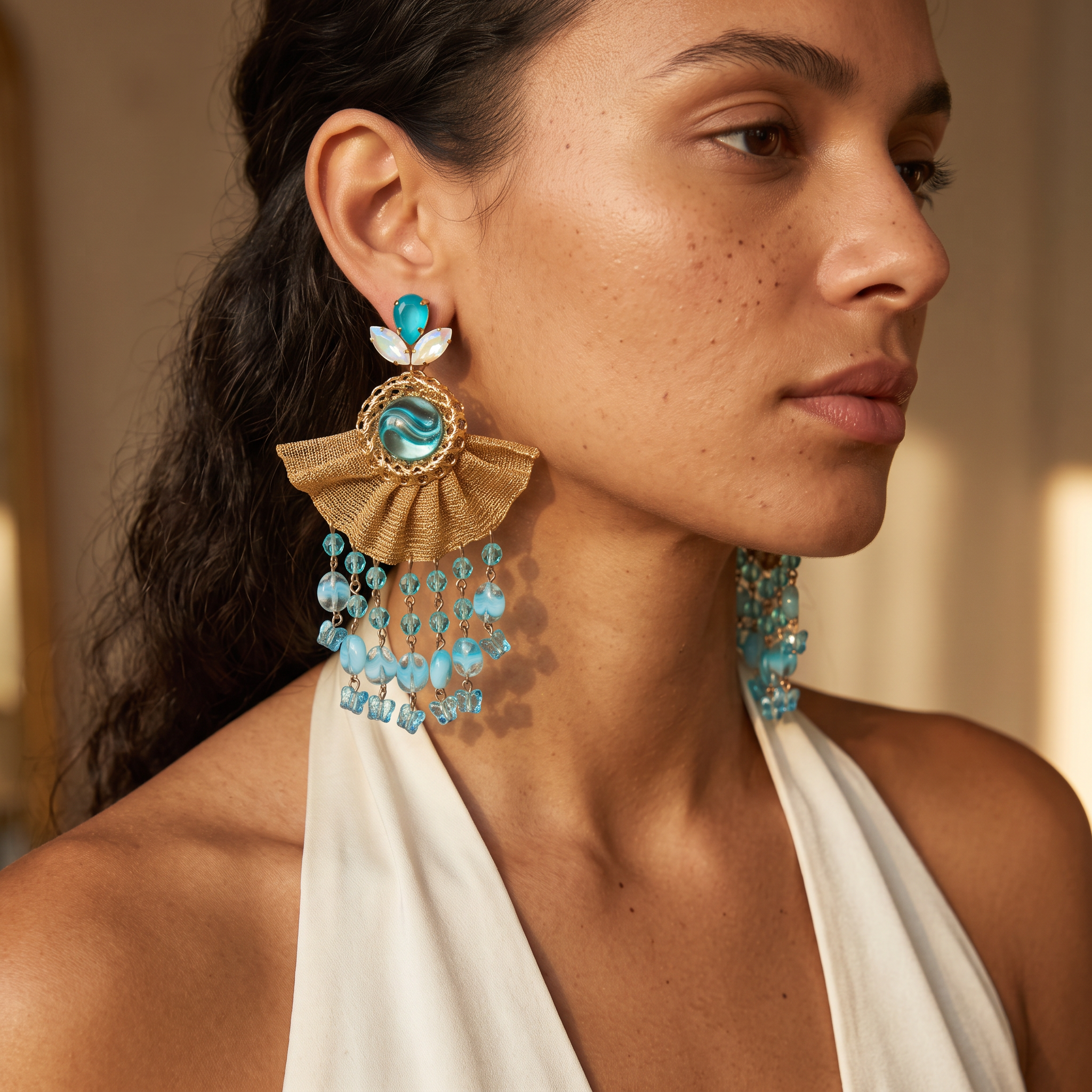 A close-up of a woman wearing a pair of dramatic statement earrings. The earrings feature a gold-toned fan-shaped metal frame with a turquoise blue stone at the center, a small white leaf detail, and cascading strands of faceted light blue beads.