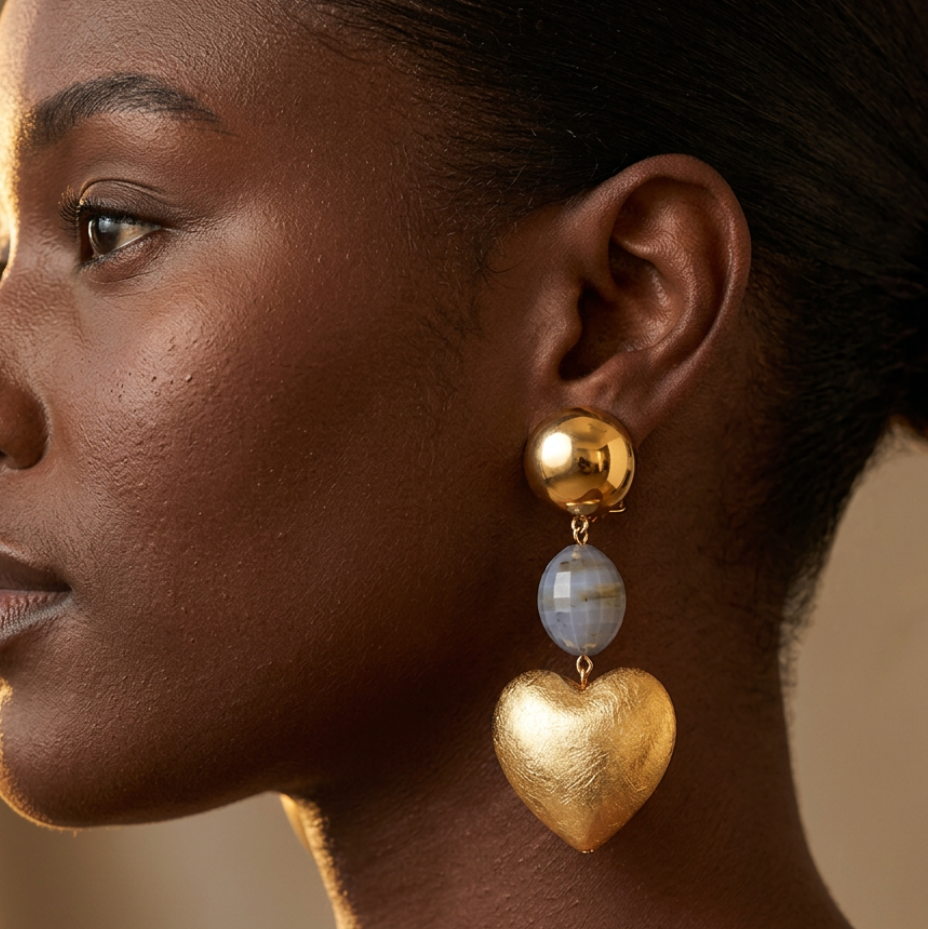 A close-up side profile of a woman wearing a gold drop earring with a round gold bead, a light blue stone bead, and a textured gold heart pendant.