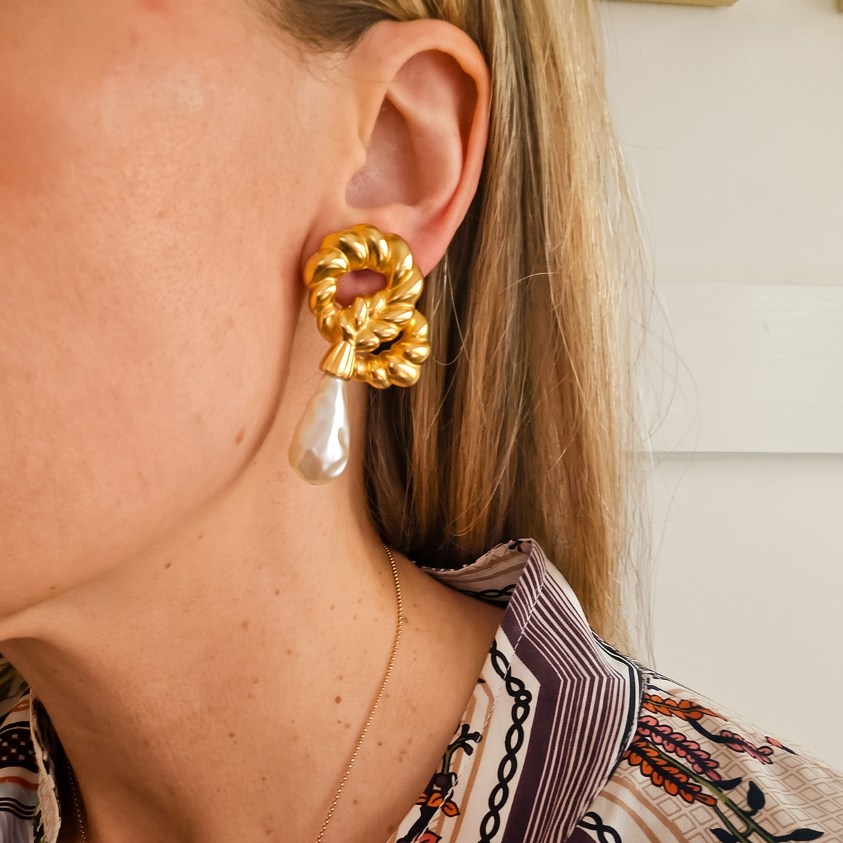 A close-up of a woman wearing a gold statement earring with a twisted rope knot design and a dangling teardrop pearl, paired with a patterned blouse.
