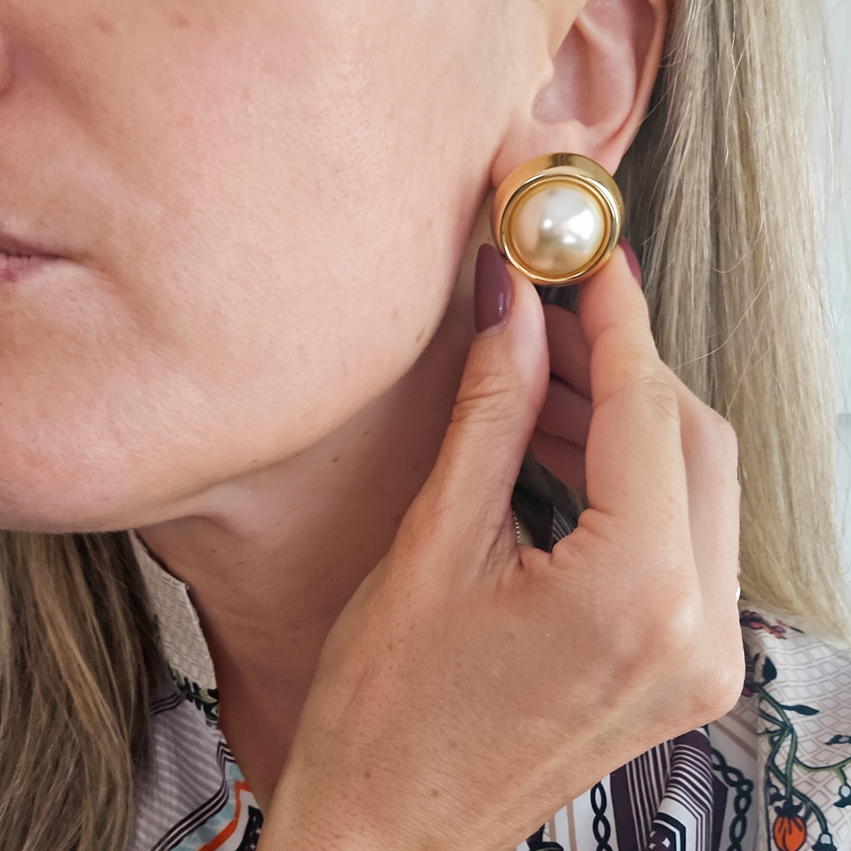 A close-up of a woman wearing a round gold earring with a white faux-pearl center, with her hand adjusting it.