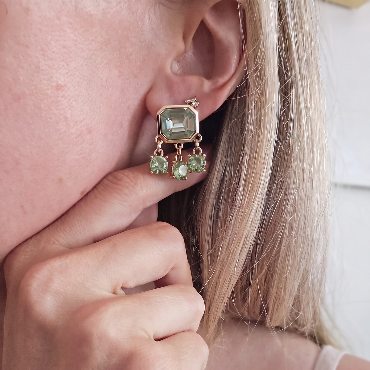 A close-up of a woman's ear wearing a gold dangle earring with a rectangular green gemstone and two smaller green hanging stones.